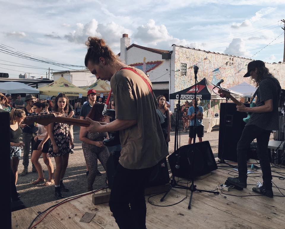 Life at CCAD, 934 Gallery in Milo Grogan District, Image of two male musicians playing guitar on a wooden stage overlooking small crowd, event marquees, and urban buildings in the background