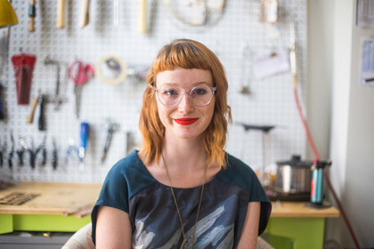 Fashion Design,  Photo of Alumni Maddie Etter of Phyllis & Hazel in front of background of hanging tools and a workspace