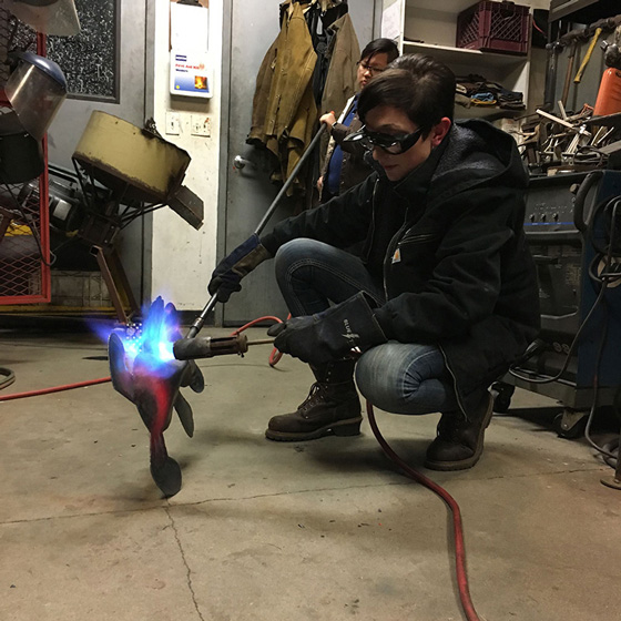 Fine Arts, Image of Grace kneeling on floor metal working with torch and metal teddy bear with metal working machinery in background