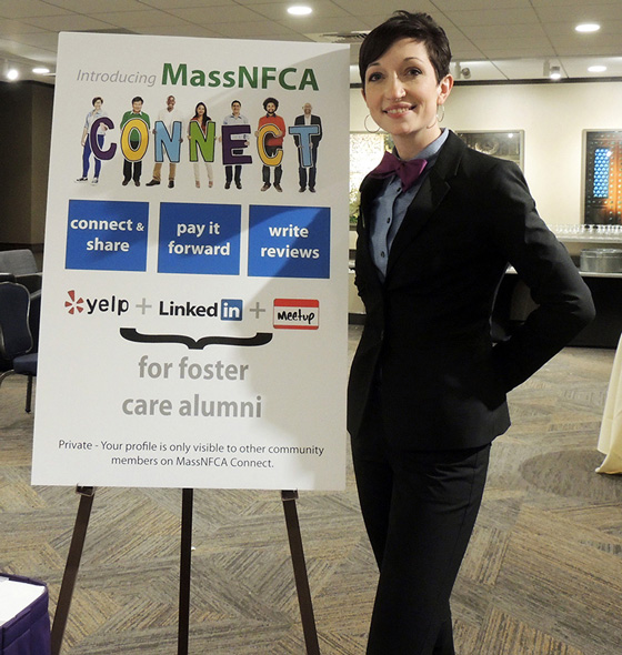 Fine Arts, Image of Grace in black suit over blue shirt with purple bow tie standing next to sign Introducing "MassNFCA"