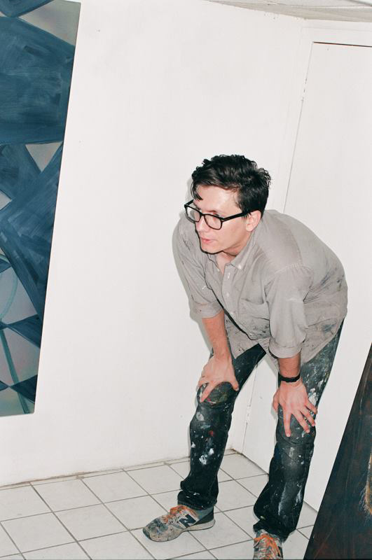 Fine Arts, Photo of Clinton leaning over with hands on knees in paint covered clothing against a white wall and white tiled floor