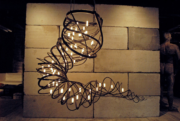 Illustration, Hanging metal swirling sculpture shaped like a tornado lit by many candles hanging within. Sculpture hangs in front of stone wall in dimly lit gallery.