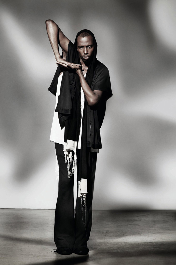 Fashion Design, Model posing in black and white, asymmetrical clothing in front of gray background. Not smiling, posing with one arm up, bent with palm facing down resting on other palm with arm pointing down, palm up