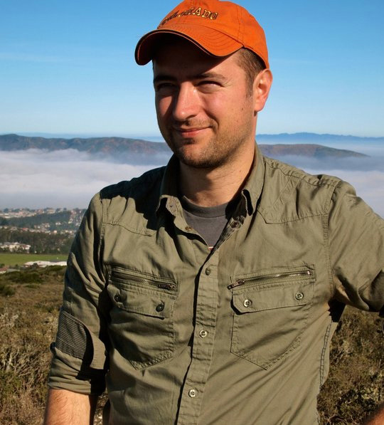 Animation, photo of Mike in a field overlooking mountains and a town. Looking slightly left of camera in an olive green button down and orange baseball cap.