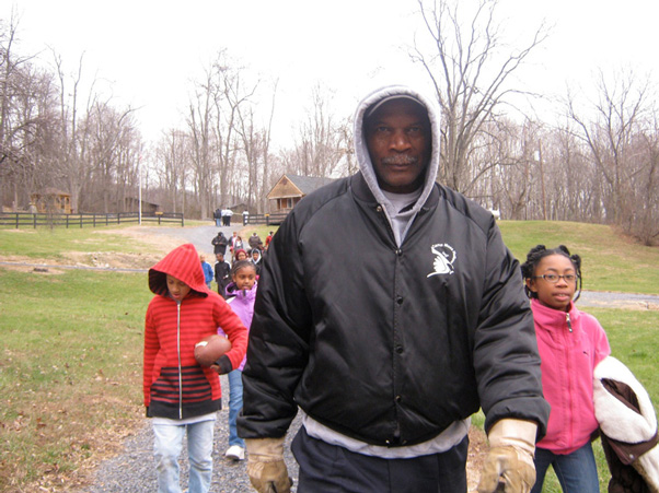 Advertising &  Graphic Design, Photo of Mike in black jacket and cloves leading a group of children down a grass lined path in fall with bare trees and gray sky in background