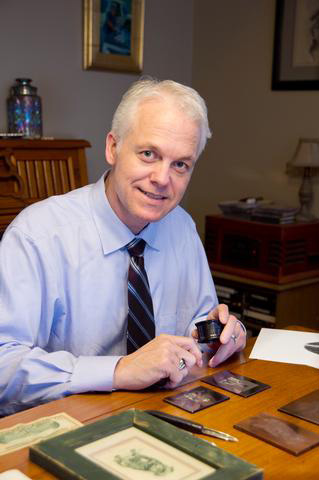 Fine Arts, image of Christopher seated at table in button down and tie smiling at camera, working with engraving tools on the kitchen table in front of hit