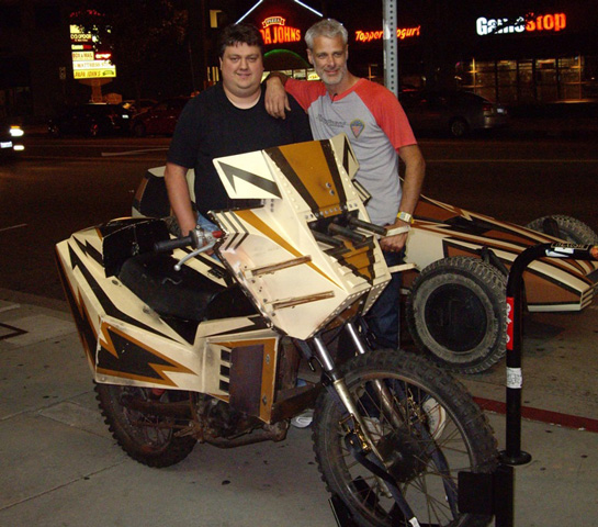 Illustration, David posing with friend behind movie prop motor cycle outside of a restaurant at night