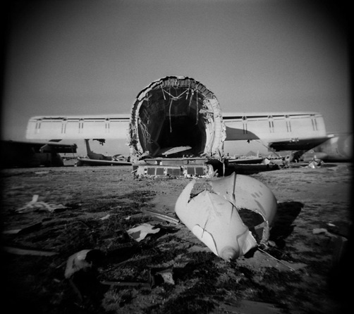 Photography, black and white image of grounded white plane with nose/cockpit ripped off lying on the ground in front of it