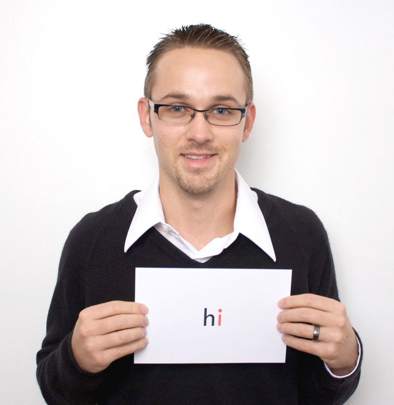 Industrial Design, Justin in black sweater poses for camera holding up a white piece of paper which reads "hi". The letter "h" is black, while the "i" is red