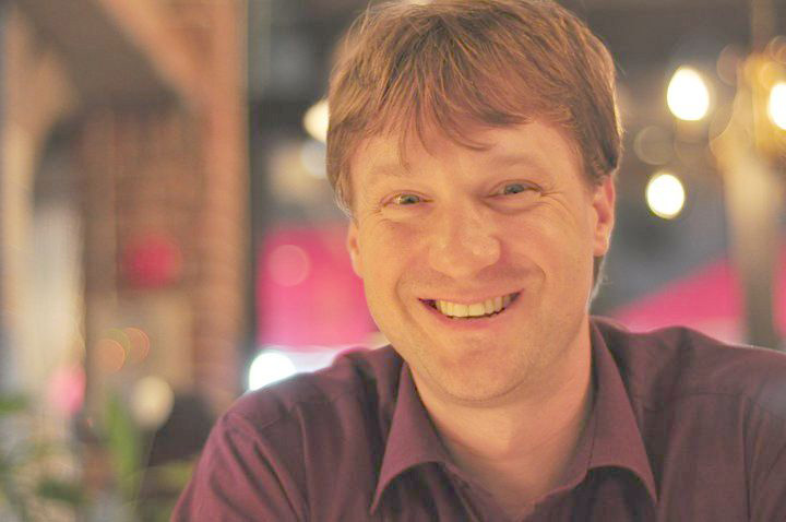 Fine Arts, Close up of Christopher Maslon in dark red polo, smiling, against a blurred night time background