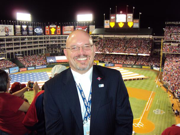 Industrial Design, Photo of Gary posing for photo, smiling, in suit standing in stands of baseball field with back to field.