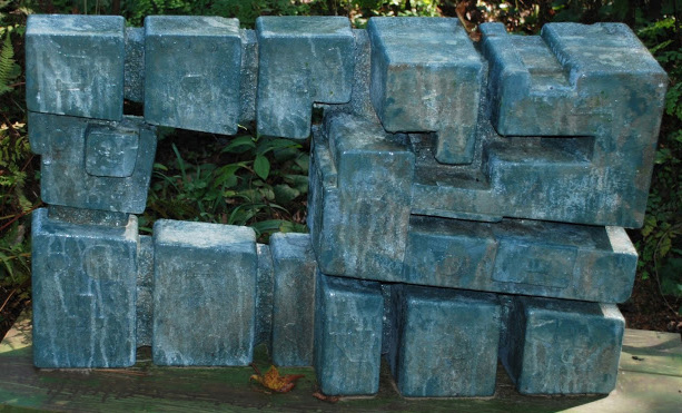 Fine Arts, Large geometric concrete sculpture with rectangle cut out center sitting in an outdoor setting with greenery in the background