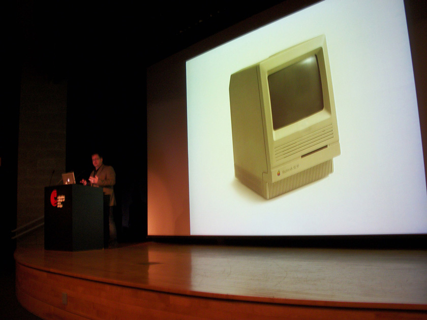 Advertising & Graphic Design, Image of presenter at podium talking and gesturing with hands. Large projector screen behind him shows image of early Macintosh computer in cream colored casing