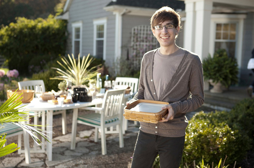 Photography, Image of Matthew smiling holding a small basket in an outdoor, backyard setting with set table, lush greenery, and gray paneled house in view.