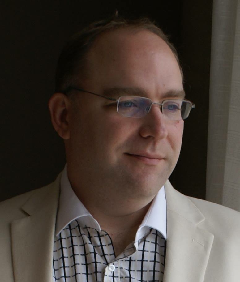 Film & Video. Headshot of Steve in cream suit jacket over black and white patterned button down, not smiling, looking away from camera into distance, dark background of a wall behind him