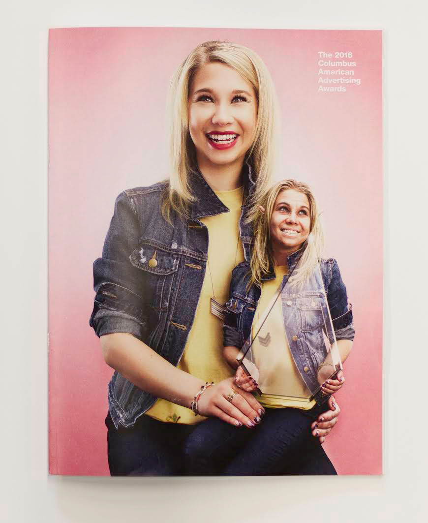 Advertising and Graphic Design, Cover of 2016 Addy's book featuring a student holding a smaller version of themselves who is holding a glass Addy award against a pink backdrop