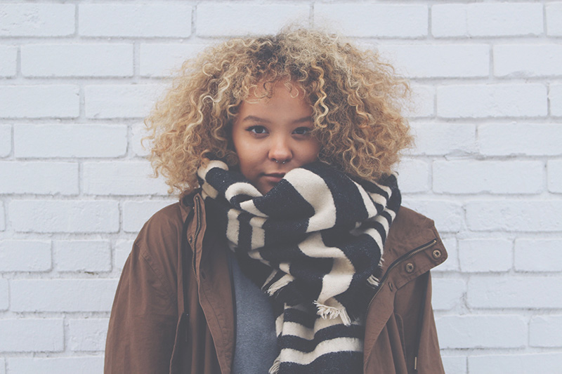 Fashion Design, Headshot of Alanta in brown coat with black and white striped scarf, facing camera not smiling, in front of white brick wall