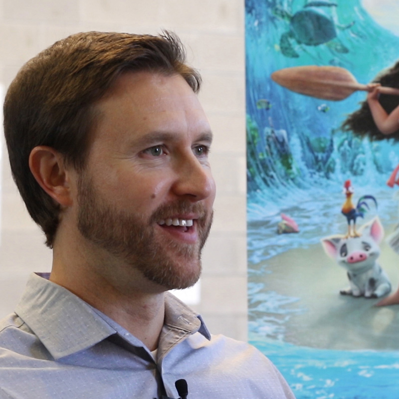 Animation, Headshot of Andy in gray button up looking slightly away from camera, with Moana poster in background