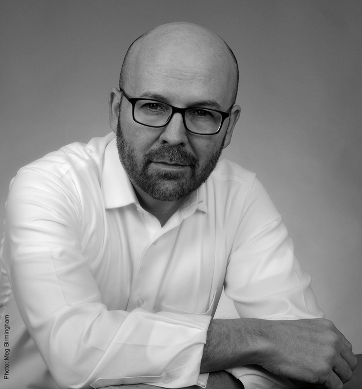 Illustration, Black and white headshot of Anthony Bakerin a white button down shirt leaning on table against a soft gray background, looking into camera but not smiling