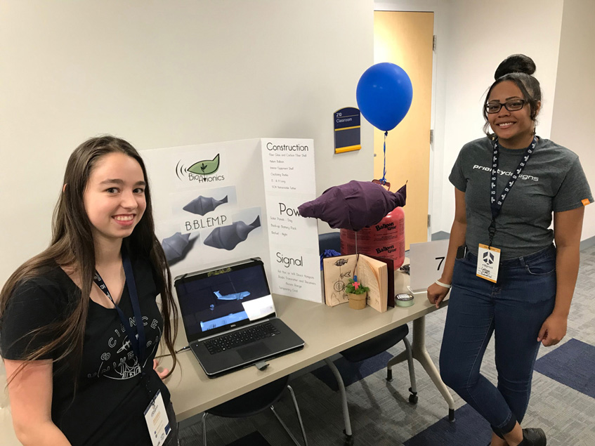 Life at CCAD, two students stand in front of table with images, 3D models, and computer models of blimp concept