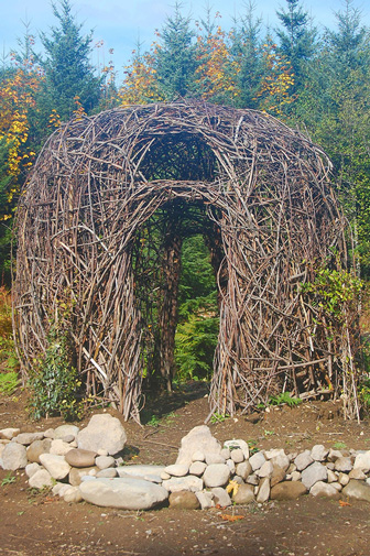 Fine Arts, Small hut made out of sticks and branches resting on dirt platform with stones around foundation against a background of trees and blue sky