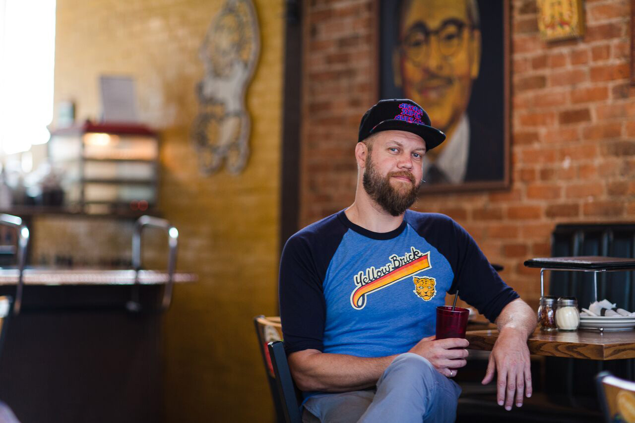 Fine Arts, Photo of CCAD Alumni Bobby Silver from Yellow Brick Pizza sitting at restaurant table holding drink cup facing camera, against blurred background of restaurant interior.