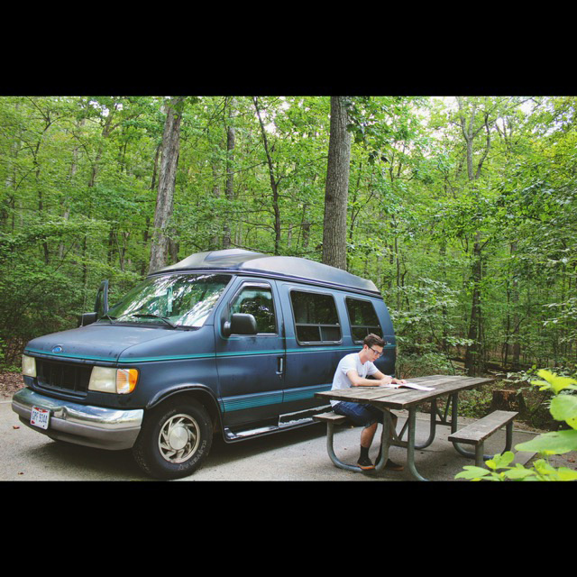 Illustration, Logan sitting at picnic table next to blue camper van amidst lush green forest of trees