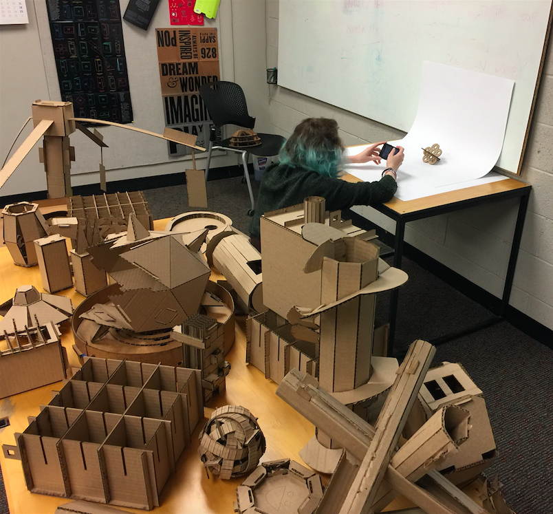 Animation,  student photographs small cardboard piece while surrounded by larger cardboard structures on a nearby table