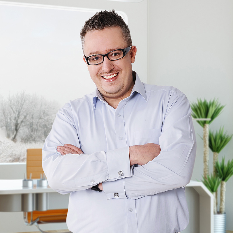 Industrial Design, Image of Devin in white button down with arms crossed smiling at camera in front of background of modern office with windows looking out on shrubbery