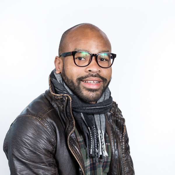 Industrial Design, Headshot of Delano Ellis, facing camera, wearing black leather jacket and plaid scarf over a green plaid shirt