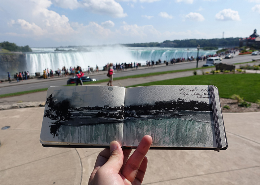 Animation, Image of hand holding up sketch of waterfall scene in sketchbook, the actual waterfall from the sketch is visible in the background behind the held up sketch book.