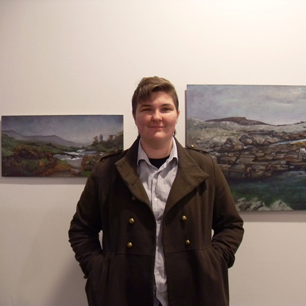 Fine Arts, Photo of Gabby in front of gallery paintings, smiling with hands in pockets of black coat layered over white button up