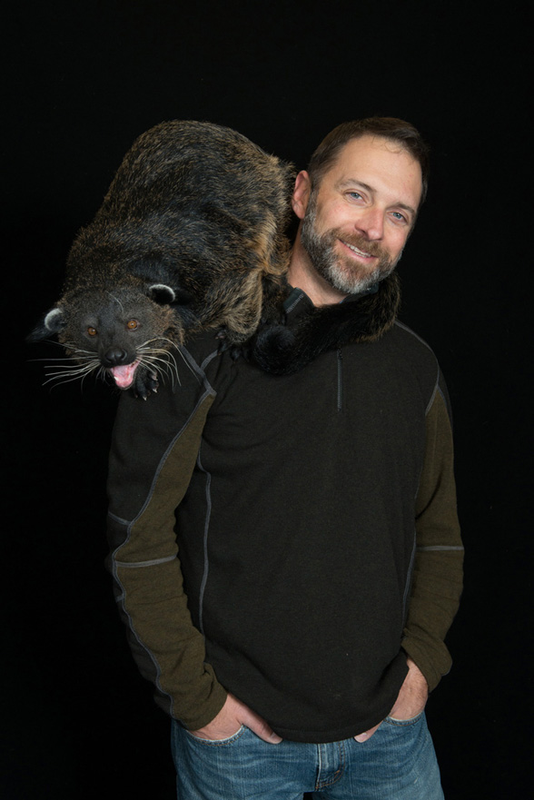 Photography, Image of CCAD grad Grahm Jones with hands in pockets and binturong (Small black, furry animal) on his shoulder in front of a black background.