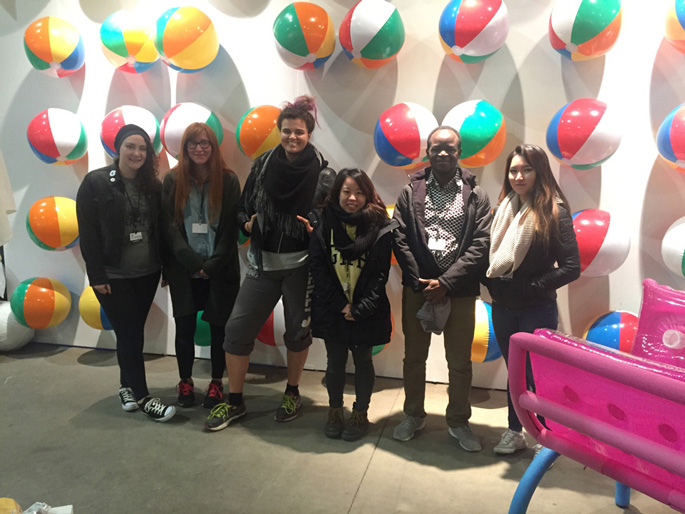 Master of Fine Arts, team of six students pose in front of wall of beach balls with pool float lounge chair in corner