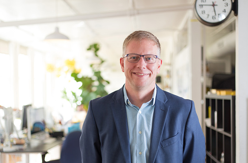 Advertising & Graphic Design, Headshot of blue suit over blue button up shirt, smiling, against background of blurred creative office setting