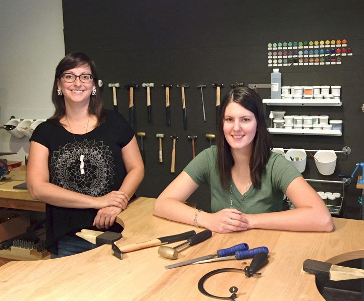 Fine Arts, Anne and Jen seated at wooden work table pose for photo against backdrop of wall of hanging craft tools and paint swatches 