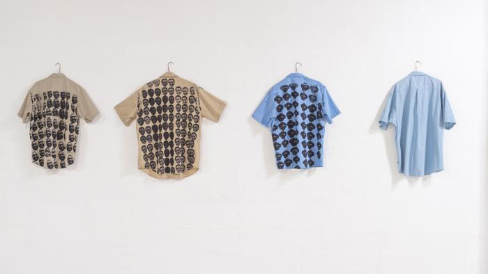 Fine Arts, four police shirts hang backwards on hangers on a gallery wall, on the backs of the shirts are printed images of African American victims of police brutality