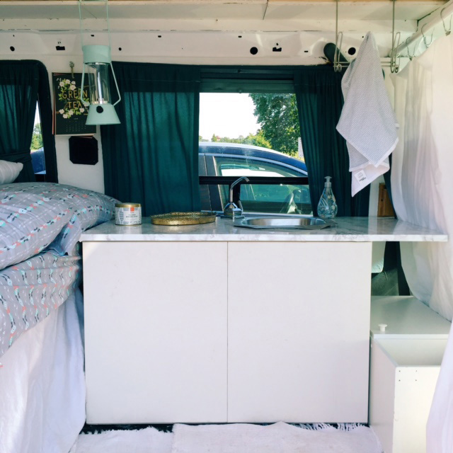 Illustration, Interior shot of converted camper van, photo focused on bedside table with sink next to edge of bed with gray sheets, mint colored lantern hangs from above, to the right a white sheet separates living space 