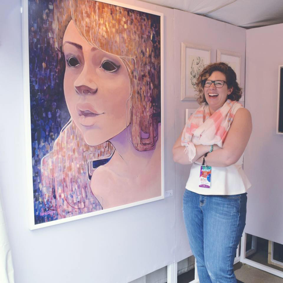 Fine Arts, Image of CCAD Alumni Lucie Shearer standing with arms crossed laughing next to painting of a woman with black eyes hanging on white gallery wall next to other work