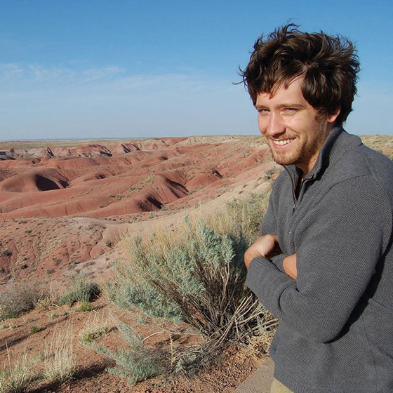 Fine Arts, Photo of Matthew smiling out at landscape with arms crossed, against a background of sparse shrubbery, hills, and desert and a blue sky