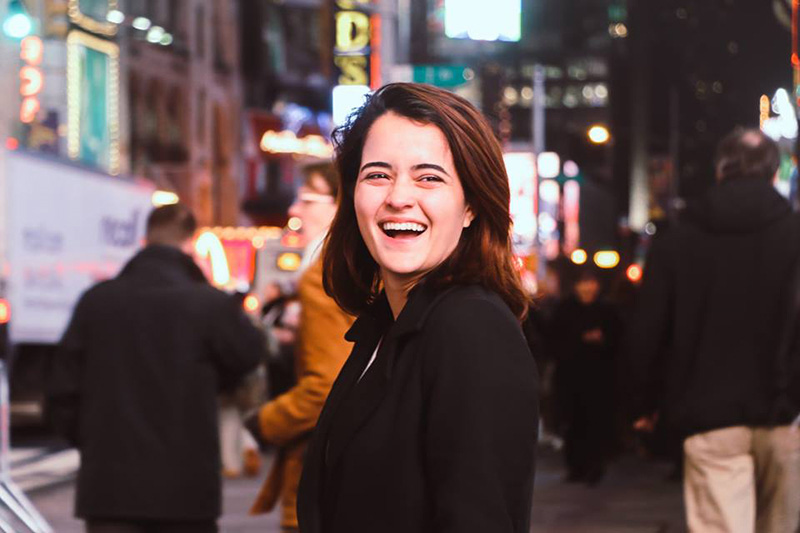 Fashion Design, Image of Natalia smiling, facing slightly away from camera with a busy street full of people in the background