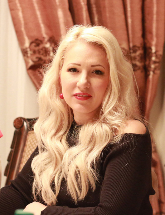 Advertising and Graphic Design, Headshot of Olga Cuzuioc wearing black shirt sitting in ornate chair against a backdrop of an ornate curtain