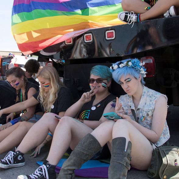 Life at CCAD, Image of students sitting behind bumper of pride parade, some on phones, other posing and smiling for photo, a pride flag hangs above their heads