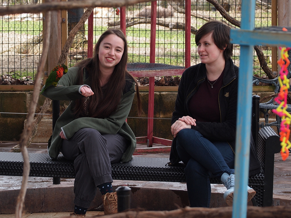 Animation, two students sit on bench in bird enclosure. One interacts with bird perched on her arm.