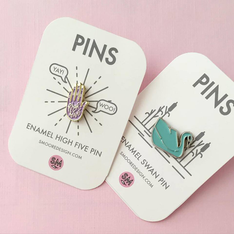 Advertising & Graphic Design, Two enamel pins on tags, to the left a pink pin of a hand says "high five", to the right is a green swan pin