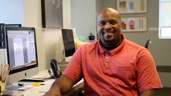 Life at CCAD, Photo of CCAD Alumni David Butler in coral polo sitting at computer desk smiling at camera