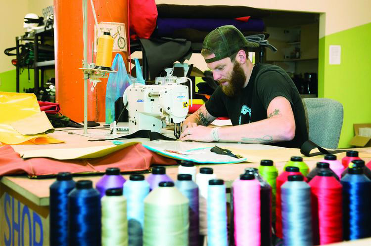 Fine Arts, Daniel McKewen from Seagull Bags working at sewing machine surrounded by colorful fabric and spools of thread in vivid blues, greens, and pinks