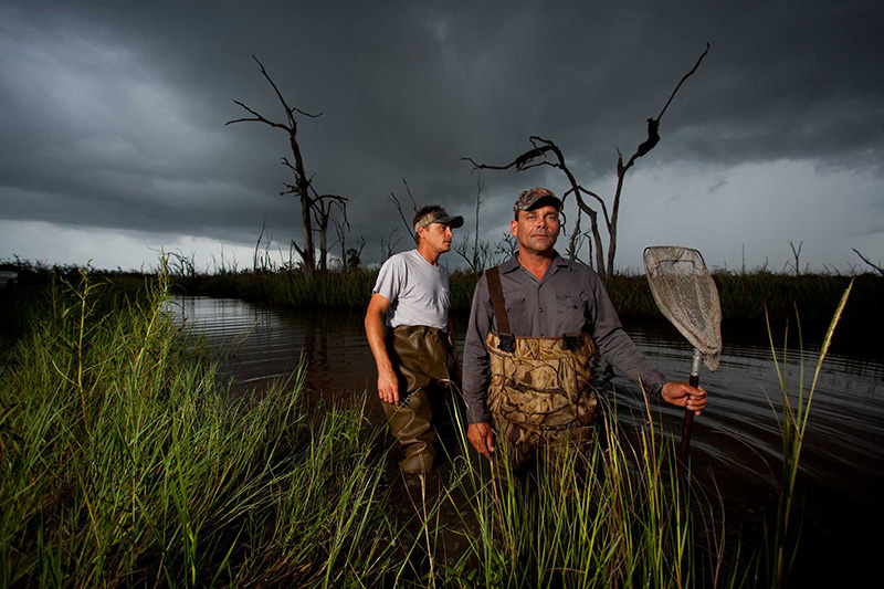 Photography, photo of two men standing in waist deep water among and tall grasses against a gray cloudy sky