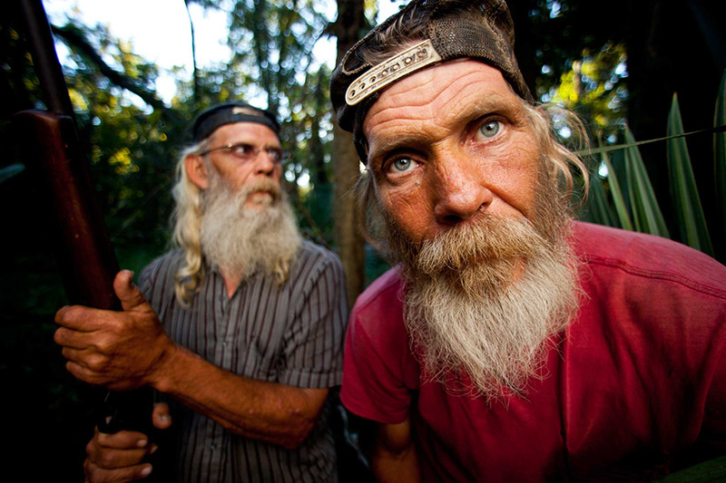 Photography, photo of two older men with beards posing for photo, one looking directly into came, one looking off into distance holding shotgun 