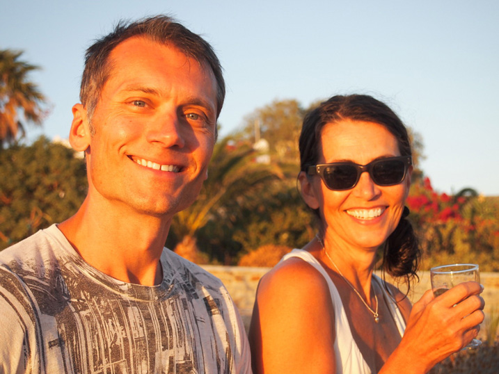 Illustration, Salli Swindell and Nate Padavick smiling for photo, Salli in sunglasses holding drink, Nate in gray and white graphic tee against a background of colorful flowering plants and blue sky at approaching sunset